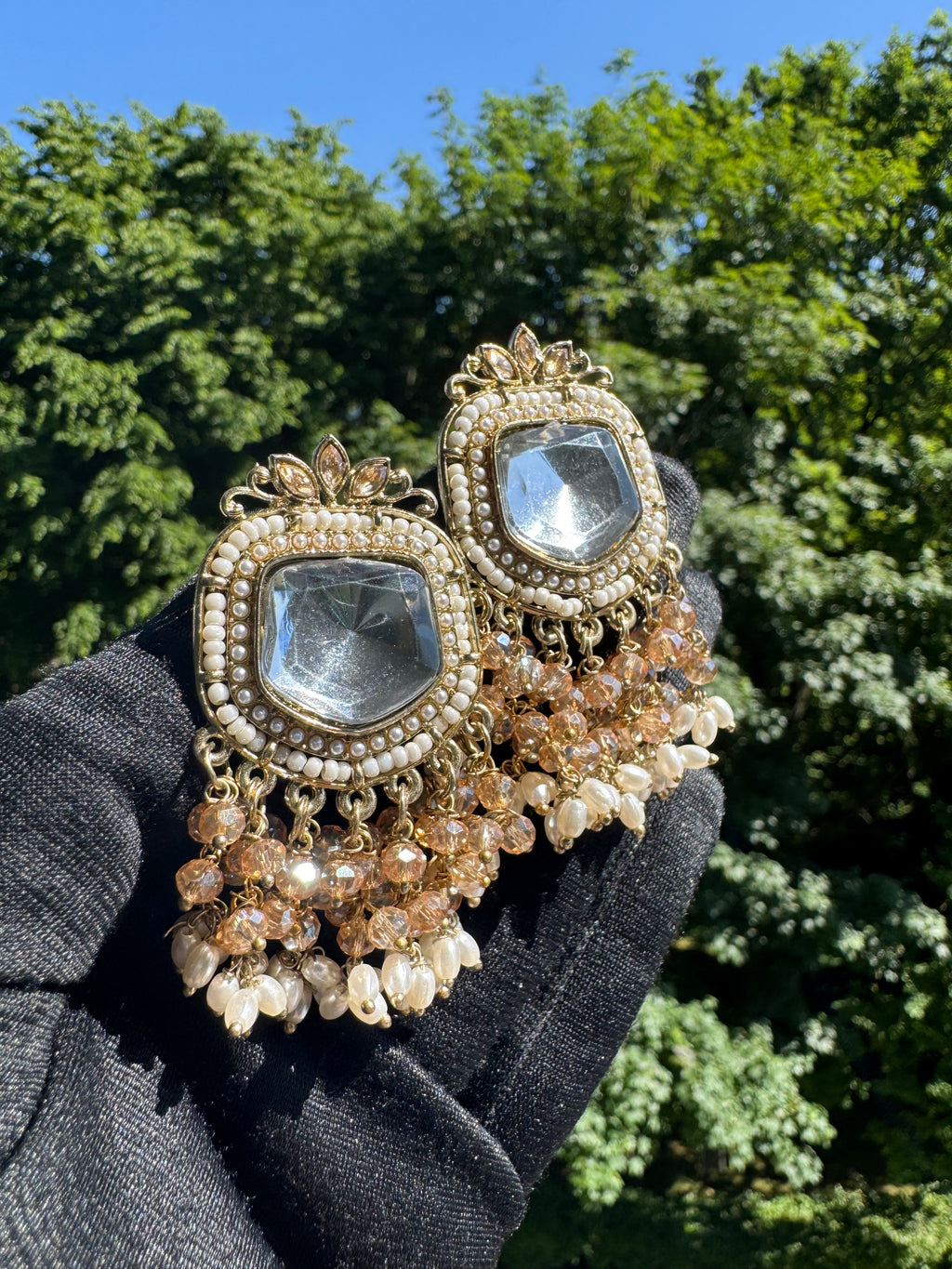 Decorative earrings with gemstones held by a gloved hand against a tree background